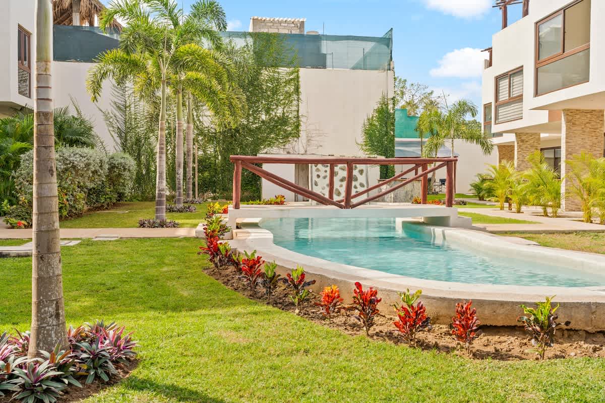 A serene outdoor scene features a long swimming pool with a rustic wooden bridge spanning its width, surrounded by a manicured green lawn and vibrant tropical foliage. Modern white buildings with brown accents are visible in the background under a bright sky.