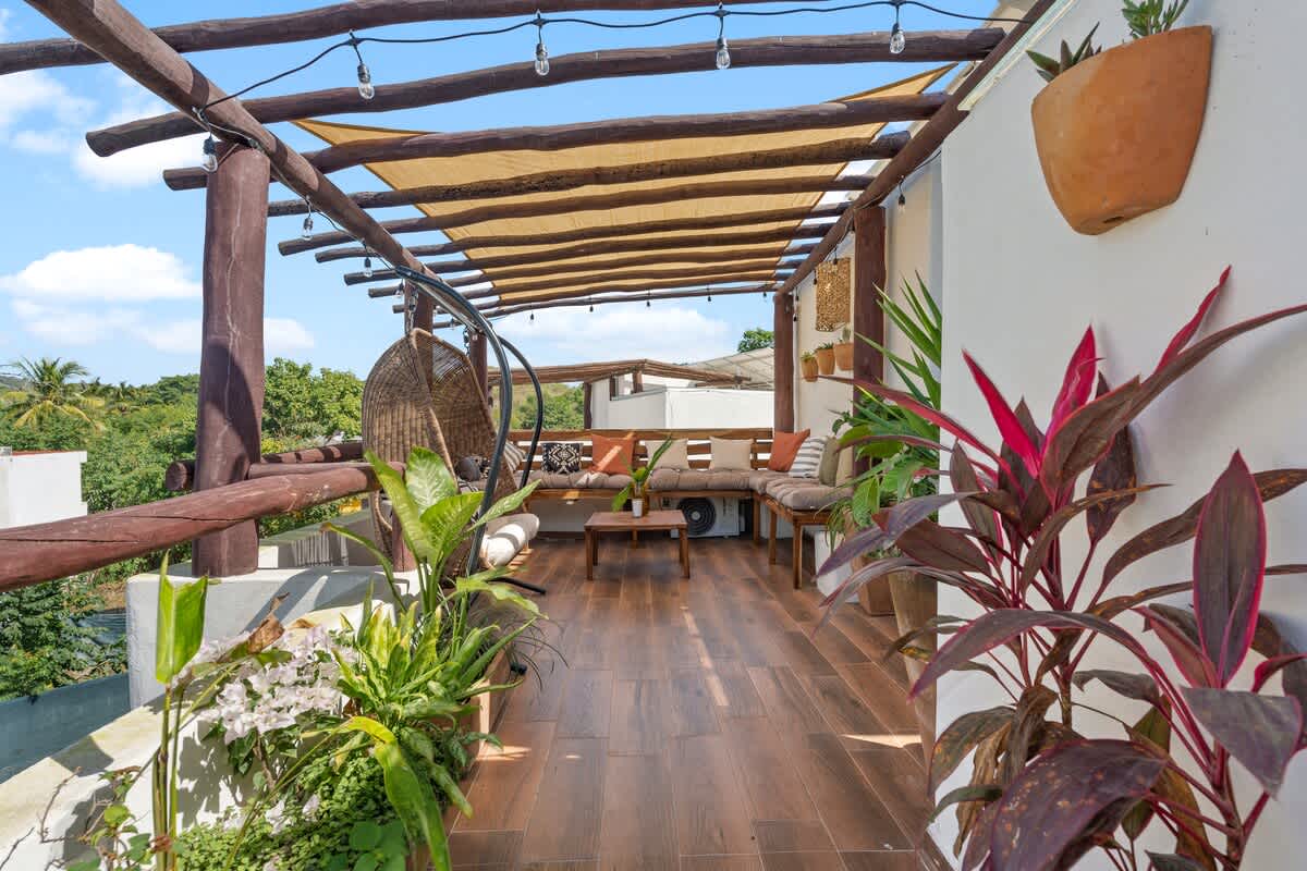An expansive rooftop deck featuring a mix of lush potted plants, a hanging swing chair, and a large L-shaped wooden bench seating area, all shaded by a pergola with string lights.