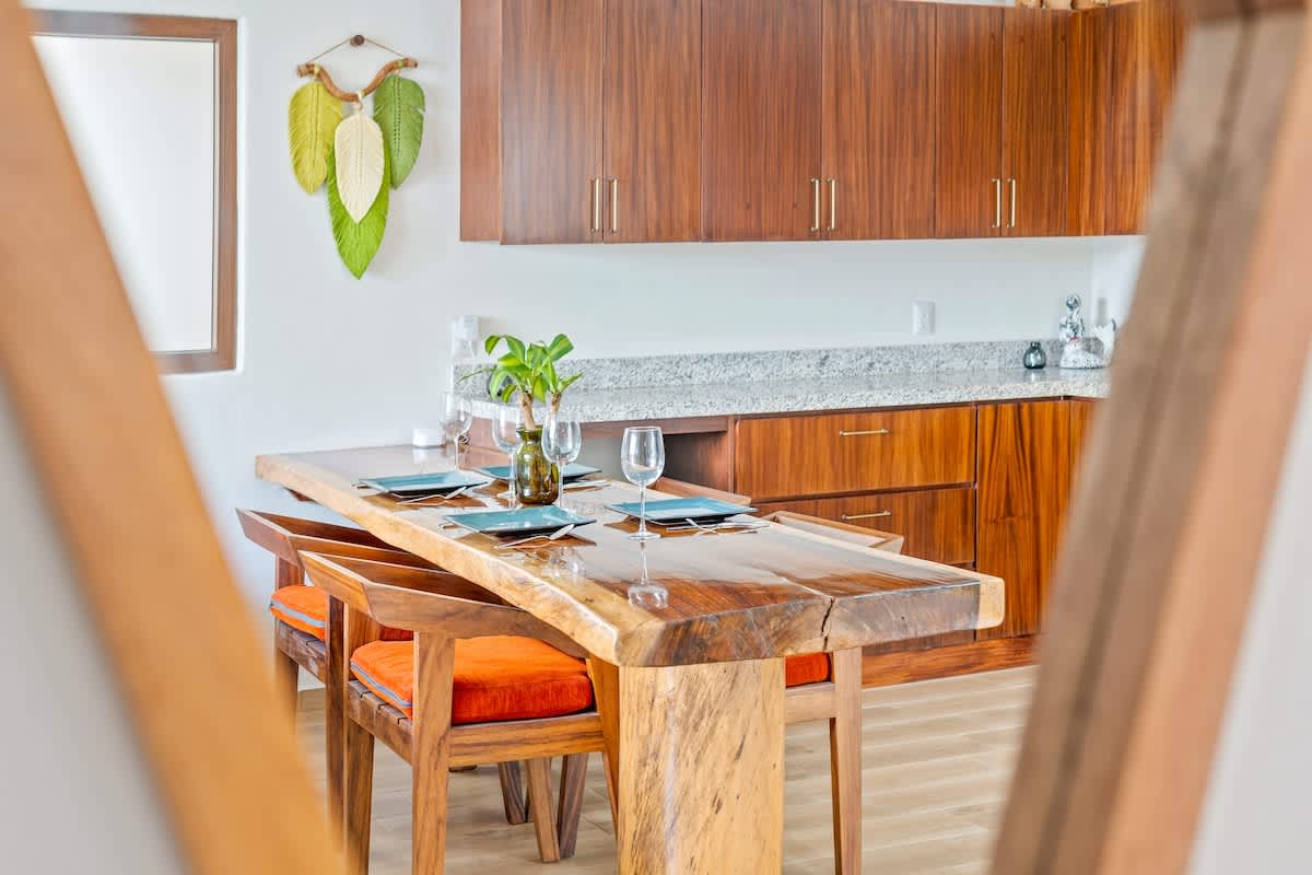 A rustic live-edge wooden dining table with orange-cushioned chairs is set with plates and wine glasses, positioned in front of a kitchen featuring dark wood cabinetry, granite countertops, and a stainless steel microwave. Decorative green leaf art hangs on the wall.