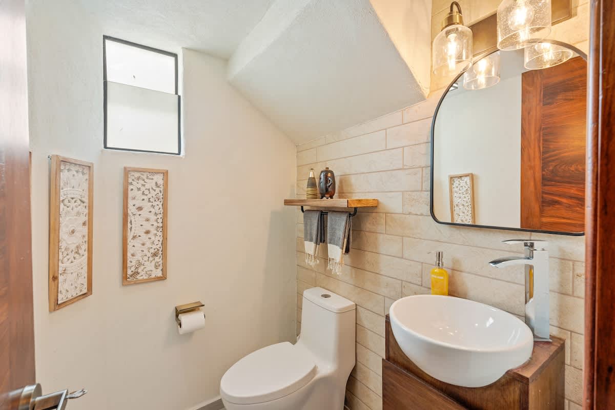 Compact bathroom with a white toilet, a vessel sink on a wooden vanity, and light-colored tiled walls. Small windows provide natural light, and decorative wall art is present.