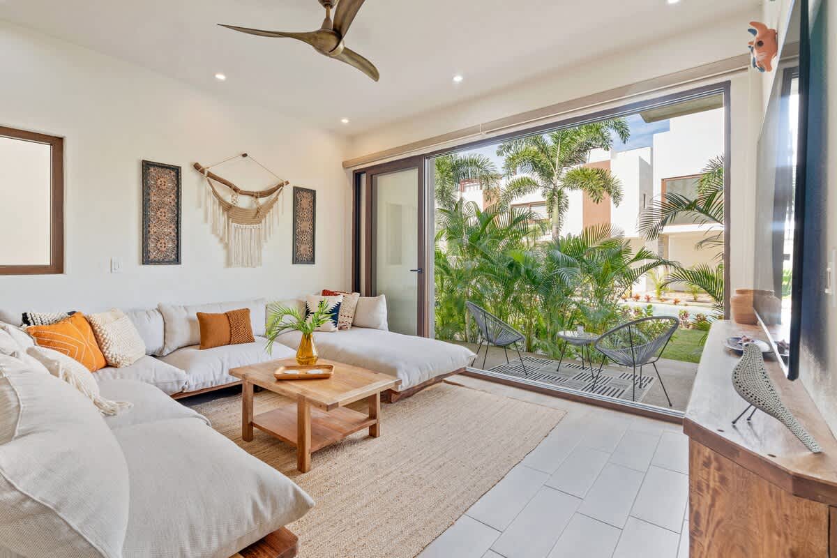 Inviting living space with a large L-shaped sectional sofa, a rustic wooden coffee table, and floor-to-ceiling sliding glass doors opening to a private patio surrounded by vibrant palm trees.