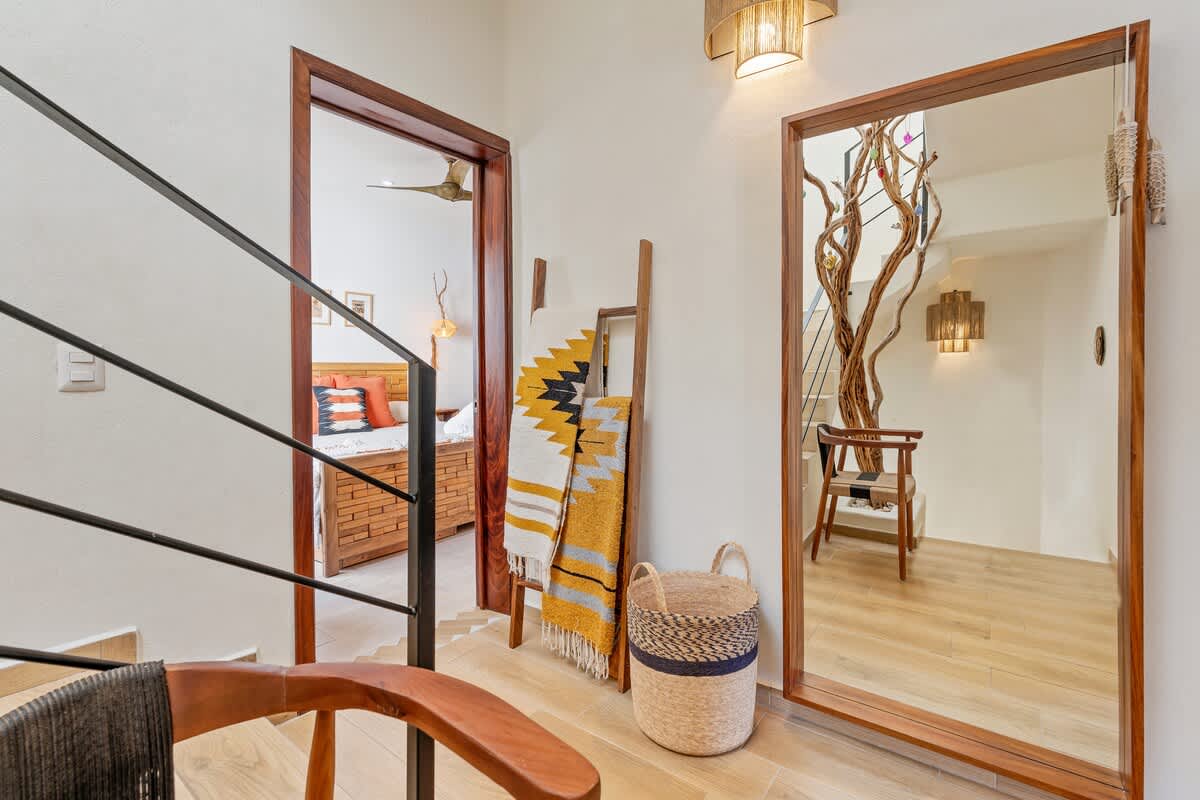 An upstairs landing features a large full-length mirror reflecting the unique tree branch and staircase from the floor below, alongside a decorative wooden ladder draped with patterned blankets. A wooden doorway opens into a bedroom, revealing a bed with a wooden headboard.