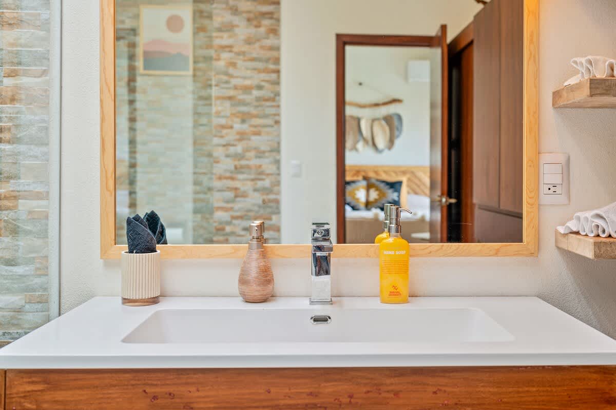 A modern bathroom vanity features a white rectangular sink with a sleek chrome faucet, a wooden-framed mirror reflecting a bedroom, and a hand soap dispenser, all against a light-colored tiled wall.