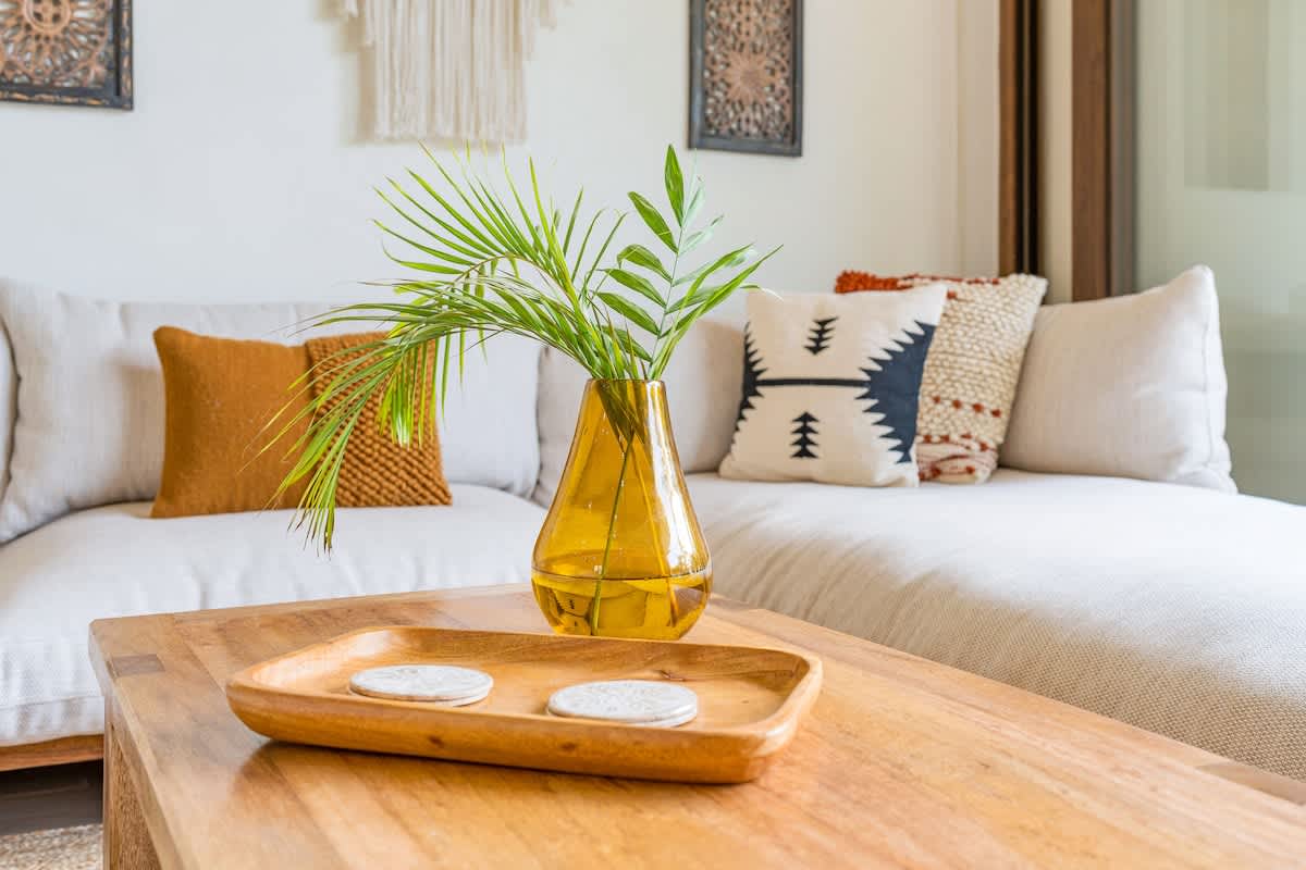A close-up highlights a rustic wooden coffee table adorned with a yellow glass vase holding green palm fronds, surrounded by a comfortable sofa with decorative pillows in earthy tones and patterns.