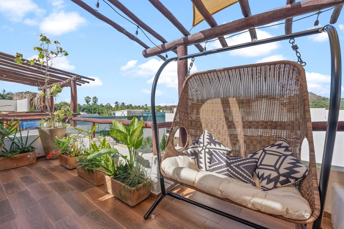 An inviting rooftop deck featuring a woven hanging swing chair with patterned pillows, surrounded by potted plants. A wooden pergola with string lights provides partial shade, overlooking distant greenery.