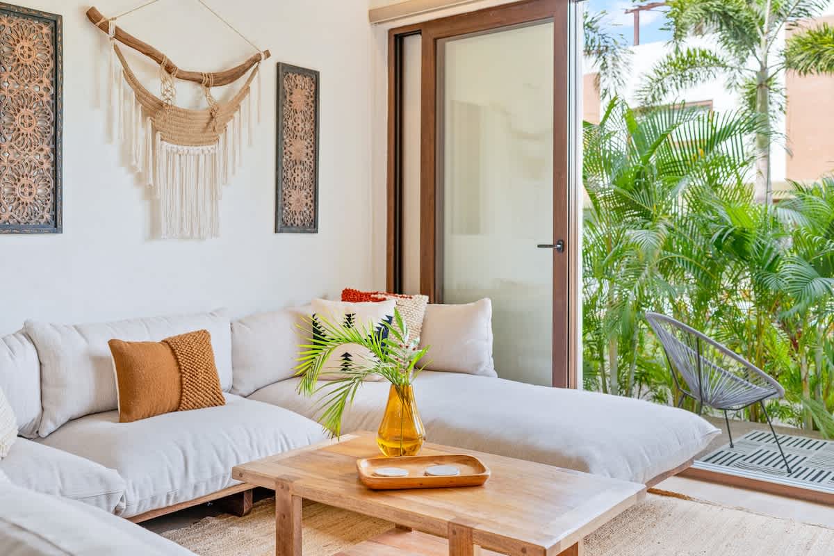 A bright living room features a large, comfortable L-shaped sofa with neutral upholstery, a rustic wooden coffee table, and a glass sliding door opening to a lush outdoor patio with tropical plants.