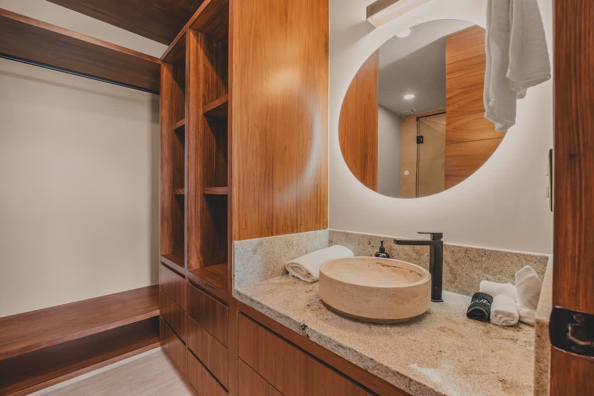 A modern bathroom vanity with a light granite countertop, a unique round stone vessel sink, and a large backlit circular mirror, adjacent to an open wooden closet with shelves and drawers.