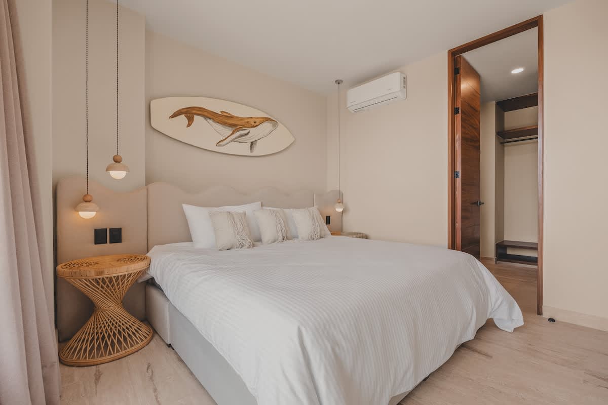 A cozy bedroom with a king-sized bed, a decorative surfboard art piece above the headboard, and an open doorway revealing a wooden closet and vanity area.