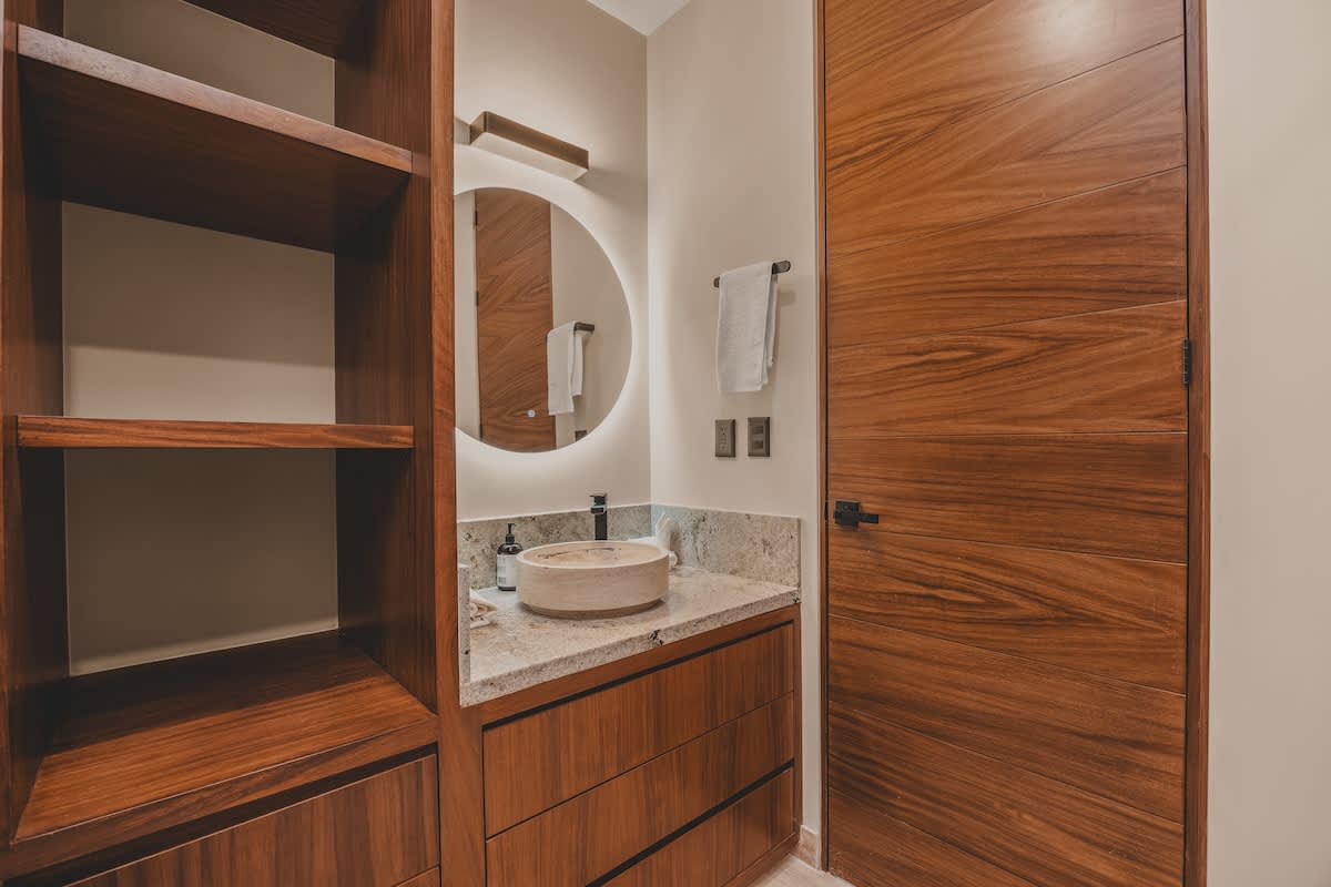 A contemporary bathroom featuring a wooden vanity with a stone vessel sink and a large backlit round mirror, next to a full-height wooden door and an open closet with shelving.