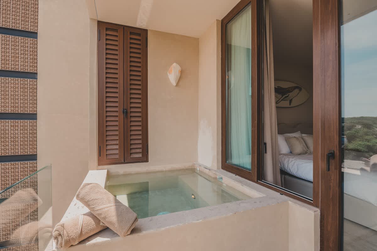 A private balcony featuring a small plunge pool or hot tub with rolled towels, adjacent to an open glass door leading into a bedroom with a bed and a surfboard wall decor.