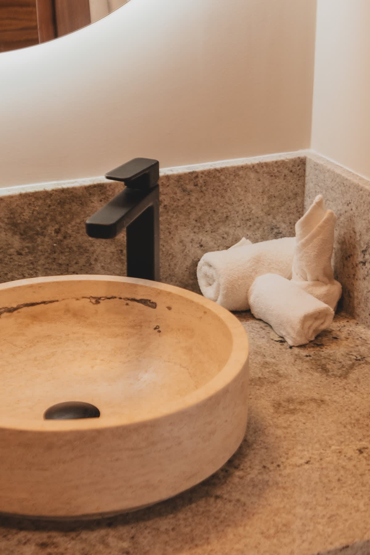 A light-colored stone vessel sink with a modern black faucet is featured on a speckled granite countertop, with neatly rolled white towels arranged beside it.