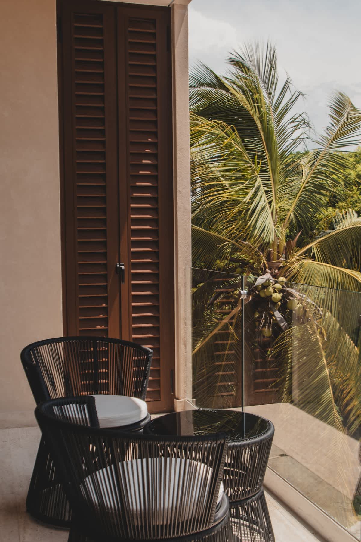 A cozy balcony features dark woven outdoor chairs and a matching round table, set against a large wooden louvered door and a glass railing, with a lush palm tree visible outside.