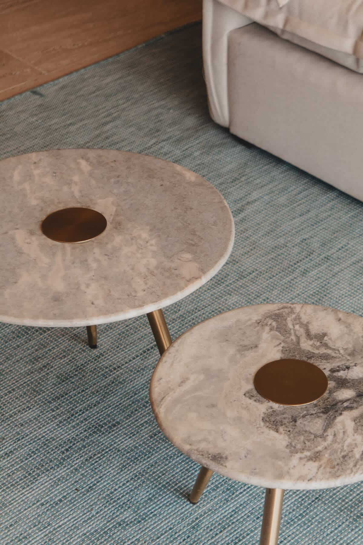 Two round, nested marble-top coffee tables with brass accents and slender gold legs sit on a textured teal-blue area rug, with a light-colored sofa partially visible in the background.