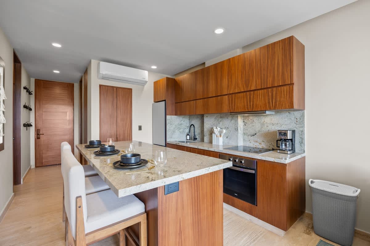 Modern kitchen and dining area with rich wood cabinetry, light granite countertops, and a matching backsplash. The dining island seats four, and the kitchen features a refrigerator, induction cooktop, and oven.