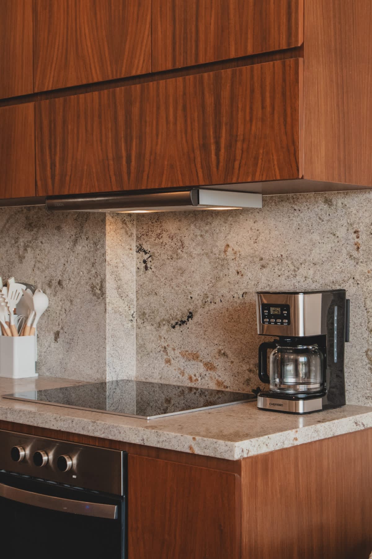 Close-up of a modern kitchen countertop featuring a sleek induction cooktop and a silver coffee maker. The light granite countertop and matching backsplash are visible, along with a white utensil holder.