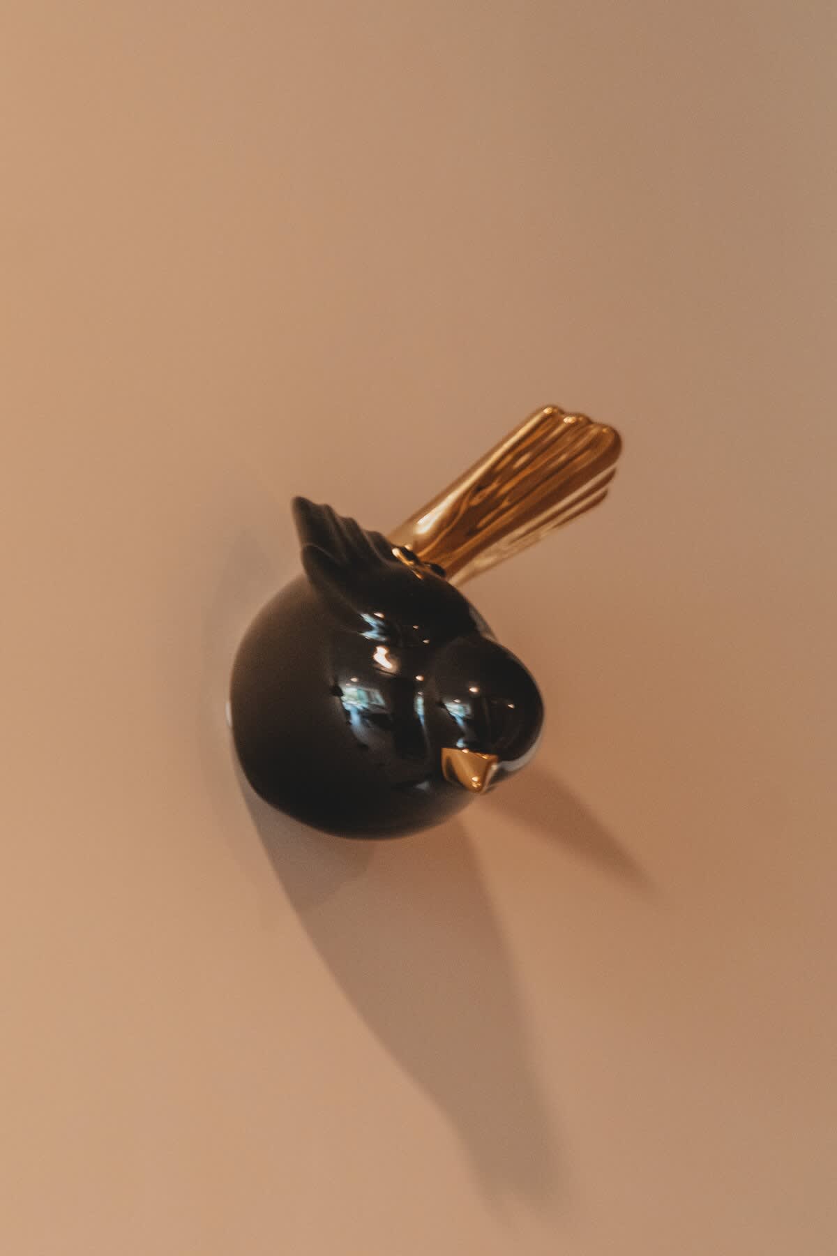 A sleek black ceramic bird figurine with a golden beak and tail feathers is mounted on a light beige wall, casting a soft shadow.
