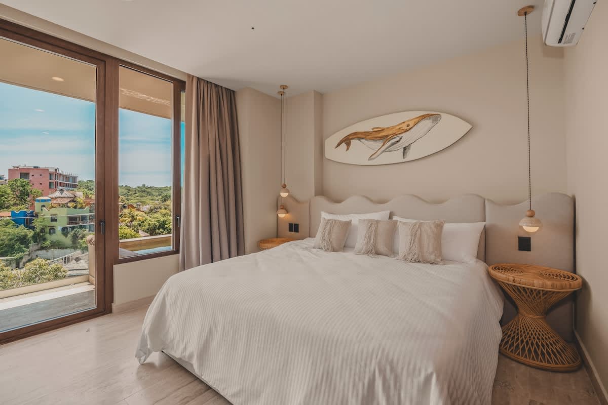 A bright bedroom featuring a king-sized bed with a light upholstered headboard, two hanging pendant lights, a unique woven side table, and large glass doors leading to a balcony with a scenic view.