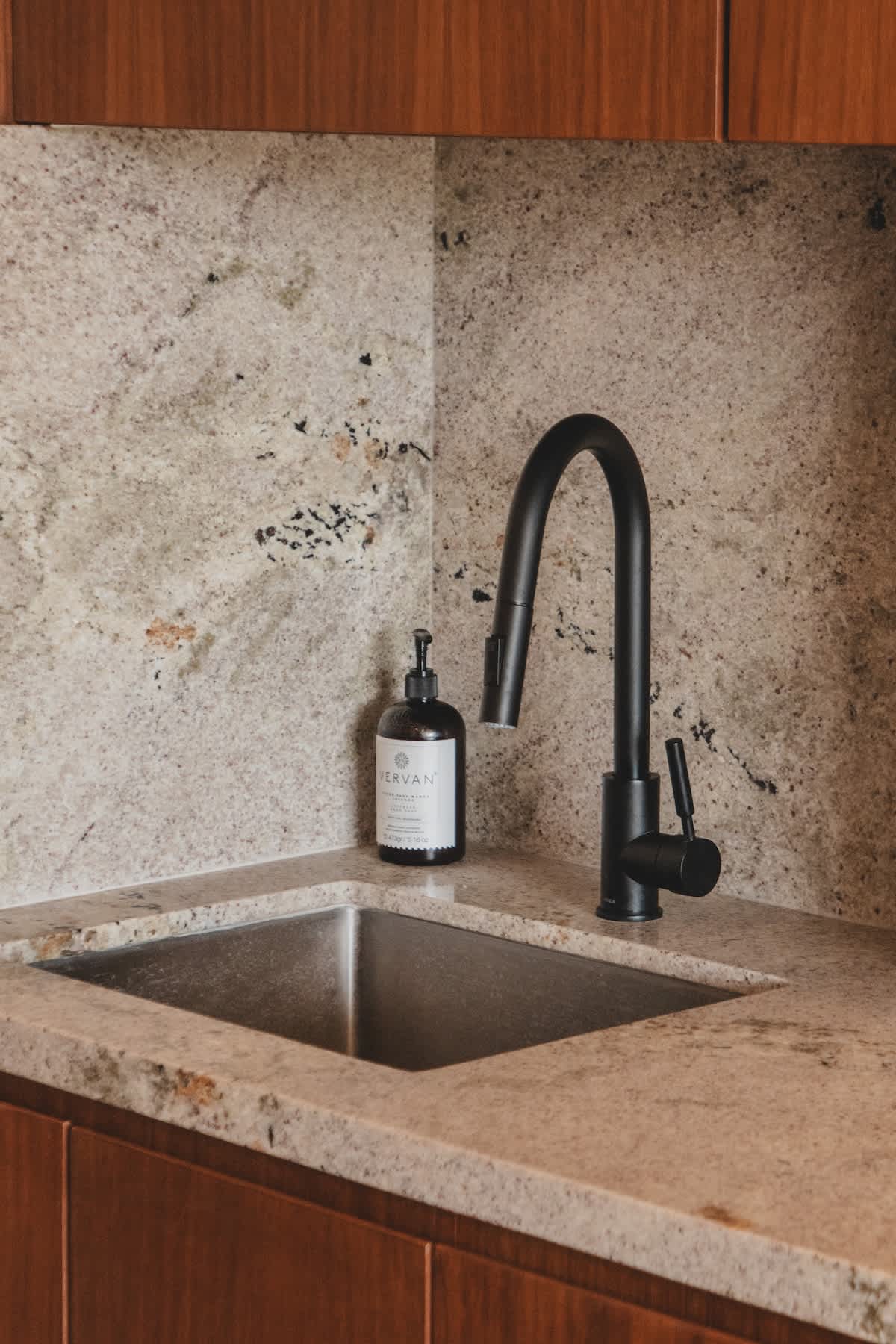 Close-up of a modern kitchen sink area with a black gooseneck faucet and a stainless steel square sink. The light granite countertop and matching backsplash are visible, along with a bottle of hand soap.