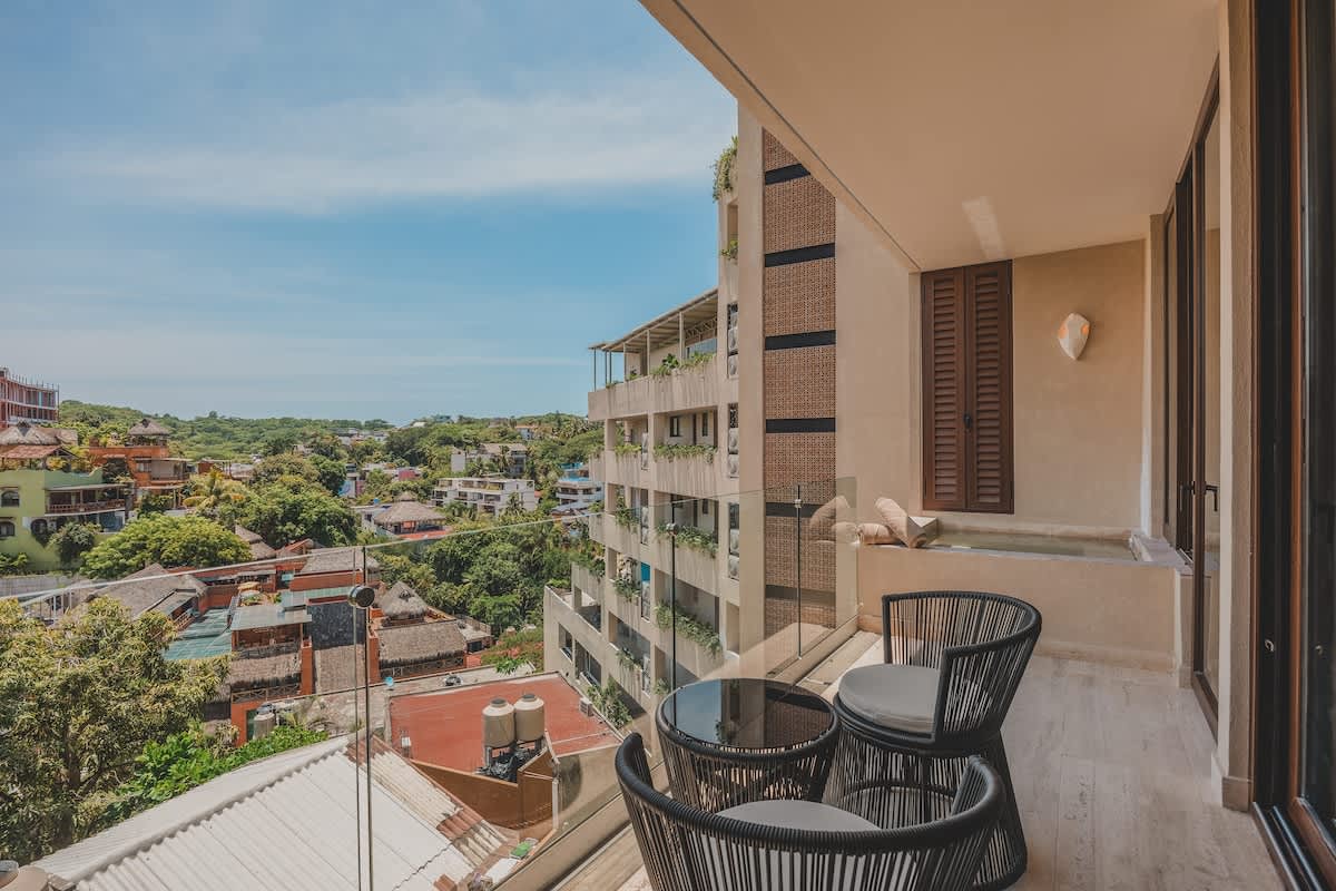 A spacious balcony with modern black woven outdoor chairs and a glass-top table, featuring a built-in plunge pool and offering panoramic views of a vibrant town and lush hills.
