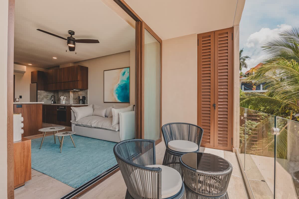 A modern balcony features two woven chairs and a round table, overlooking a lush tropical landscape, with sliding glass doors opening to a living area with a light-colored sofa and a kitchen.