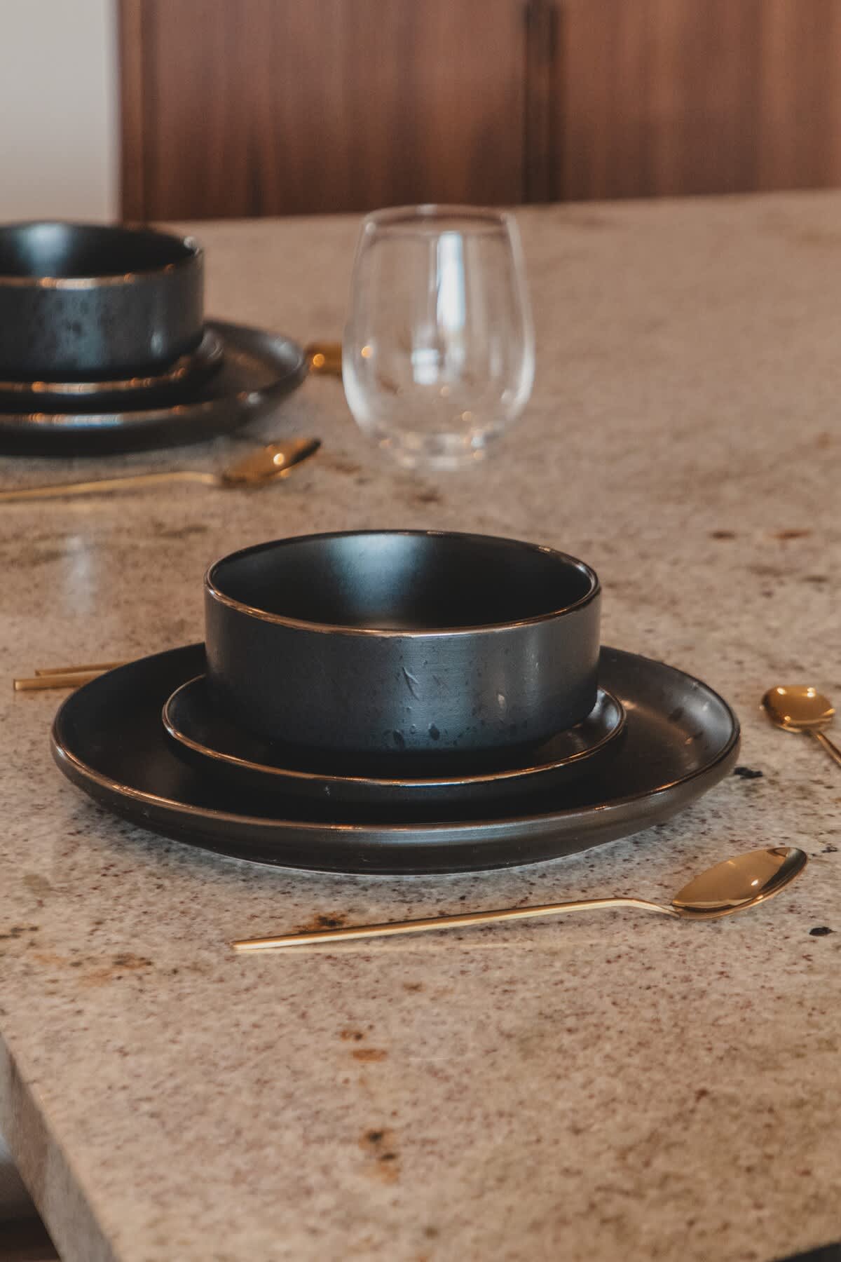 A modern place setting on a speckled granite countertop features dark, matte ceramic plates and bowls, accompanied by elegant gold spoons and a clear glass. The minimalist design suggests a sophisticated dining experience.