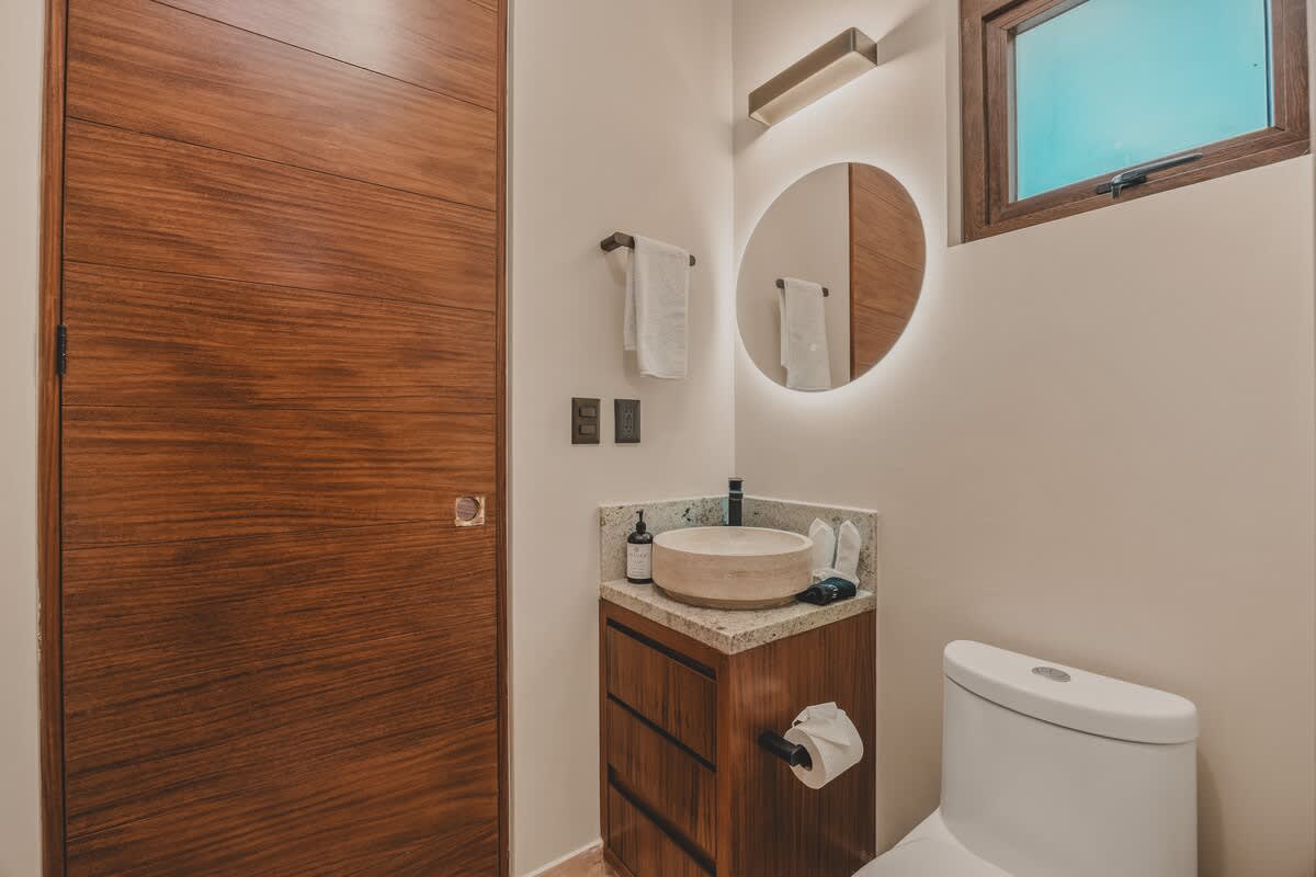 Contemporary bathroom featuring a round backlit mirror, a stone vessel sink on a dark wood vanity, a white toilet, and a sleek wooden door, with a small window above.