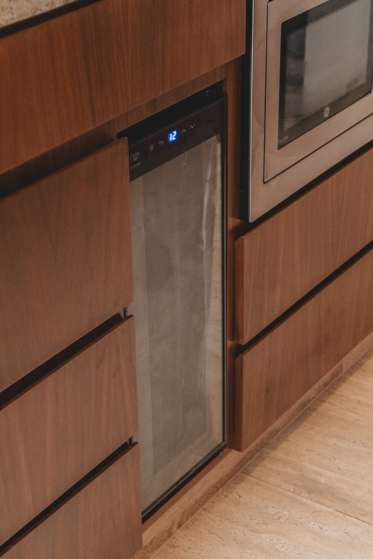 A compact, built-in wine cooler with a glass door and digital temperature display is integrated into warm wooden cabinetry, positioned next to a stainless steel microwave. This setup provides convenient beverage storage within a modern kitchen.