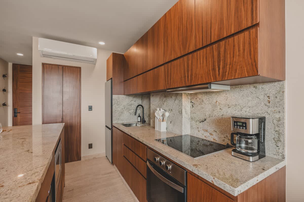 Modern kitchen with rich wood cabinetry, light granite countertops, and a matching backsplash. It features a black gooseneck faucet over a stainless steel sink, an induction cooktop, a coffee maker, and a full-size refrigerator.