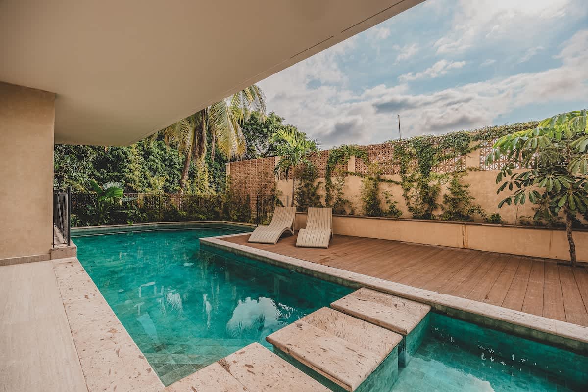 Serene swimming pool with clear turquoise water and large stepping stones, bordered by a wooden deck with lounge chairs and surrounded by vibrant tropical plants and palm trees.