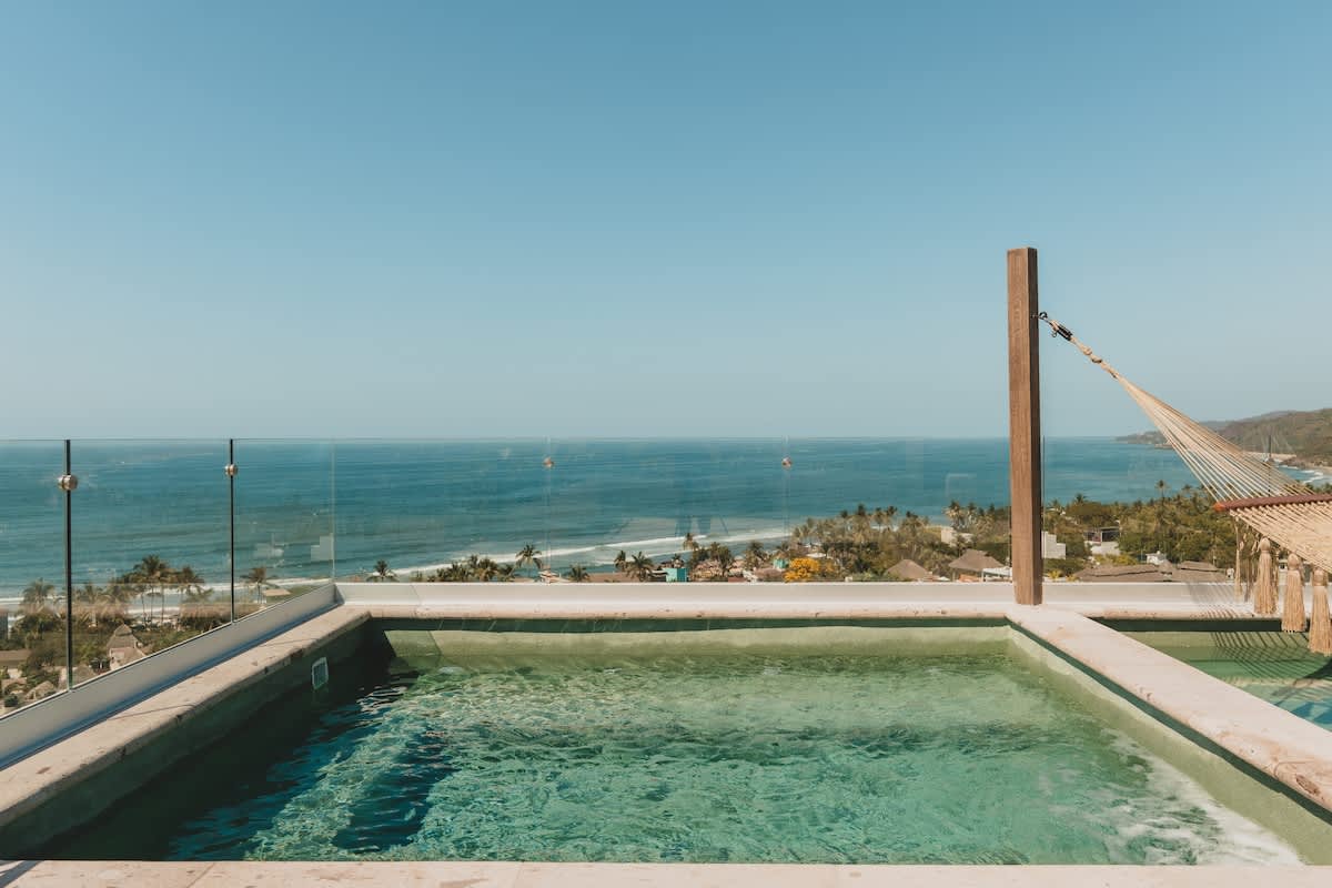 Close-up of a rooftop plunge pool with clear blue water, offering a stunning panoramic view of the ocean and distant coastline. A glass railing provides an unobstructed vista, and a hammock is visible in the background.