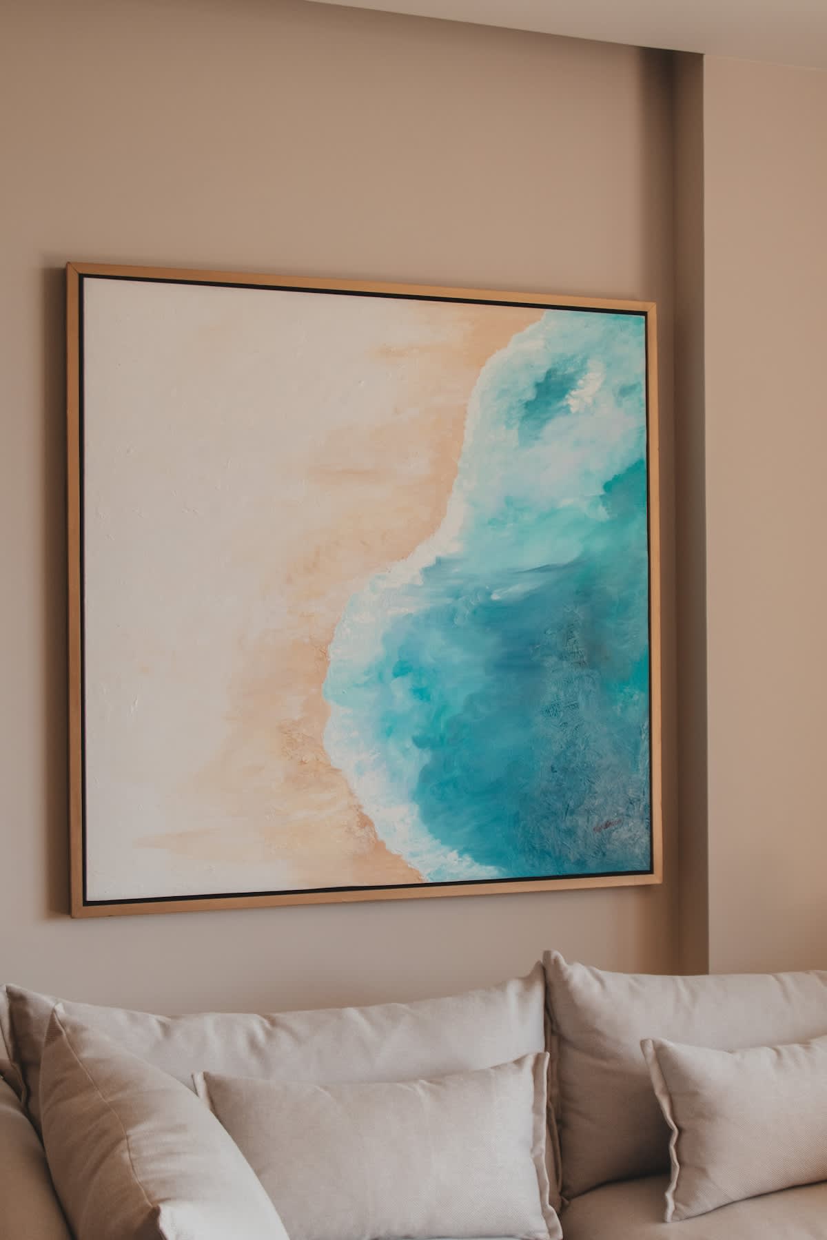 A large framed painting depicting an ocean wave breaking on a sandy beach, with vibrant blues and sandy tones, hangs above a light-colored sofa. The wall is a neutral beige.