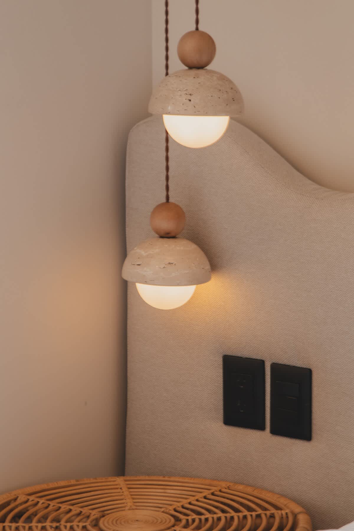 Two modern pendant lights with light-colored stone shades and wooden accents hang beside a beige upholstered headboard, illuminating the bedside area.