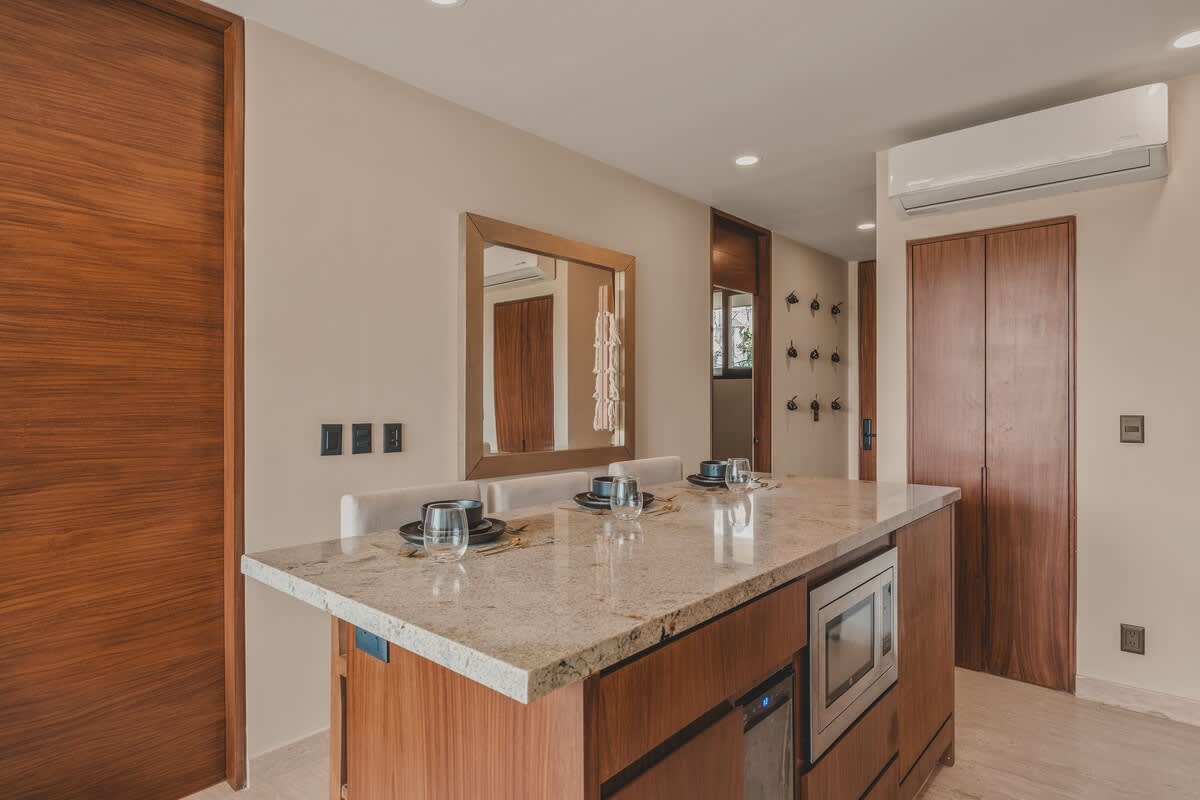 View of the dining area with a granite-topped island and seating for four, featuring an integrated microwave and wood paneling. A large mirror reflects the kitchen, and a wooden door is visible on the left.