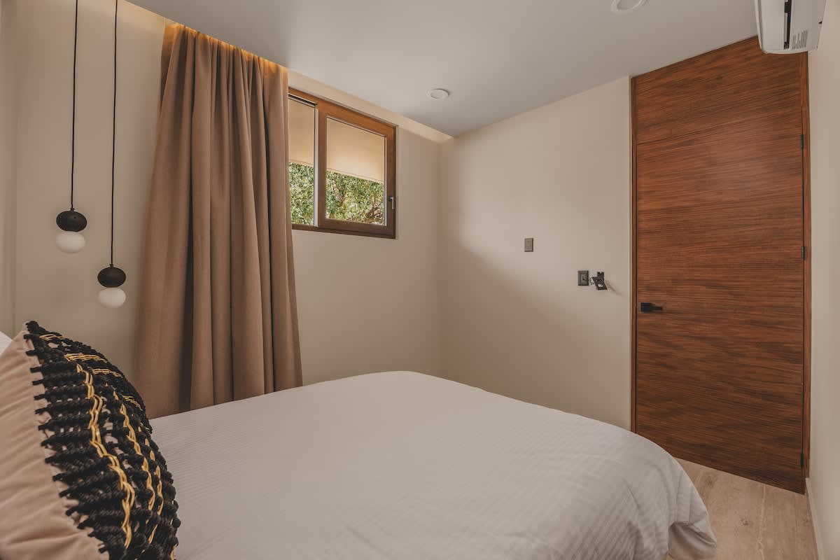 View of bedroom-2 showing a window with light-colored curtains and shades, a modern wooden door, and an air conditioning unit mounted high on the wall.