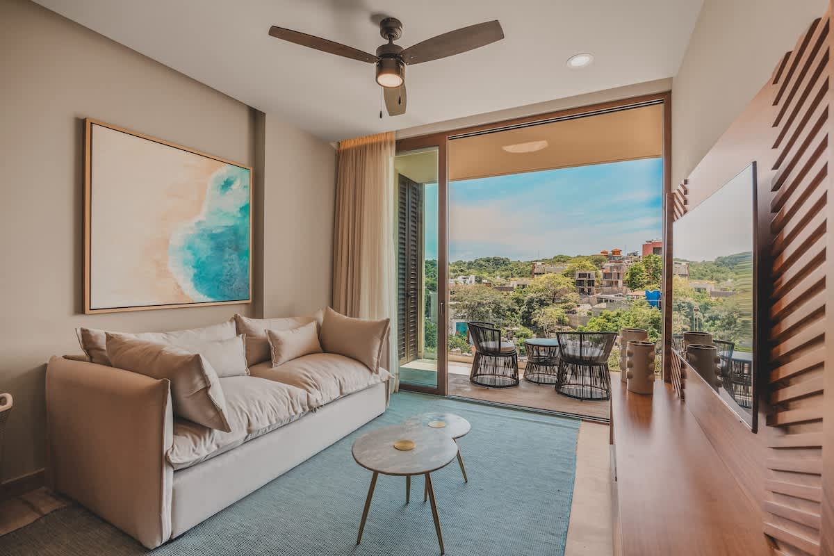 The bright living room features a comfortable light-colored sofa, a blue area rug, and a wooden media console, with large sliding glass doors opening to a balcony offering panoramic views of a lush, hilly landscape.