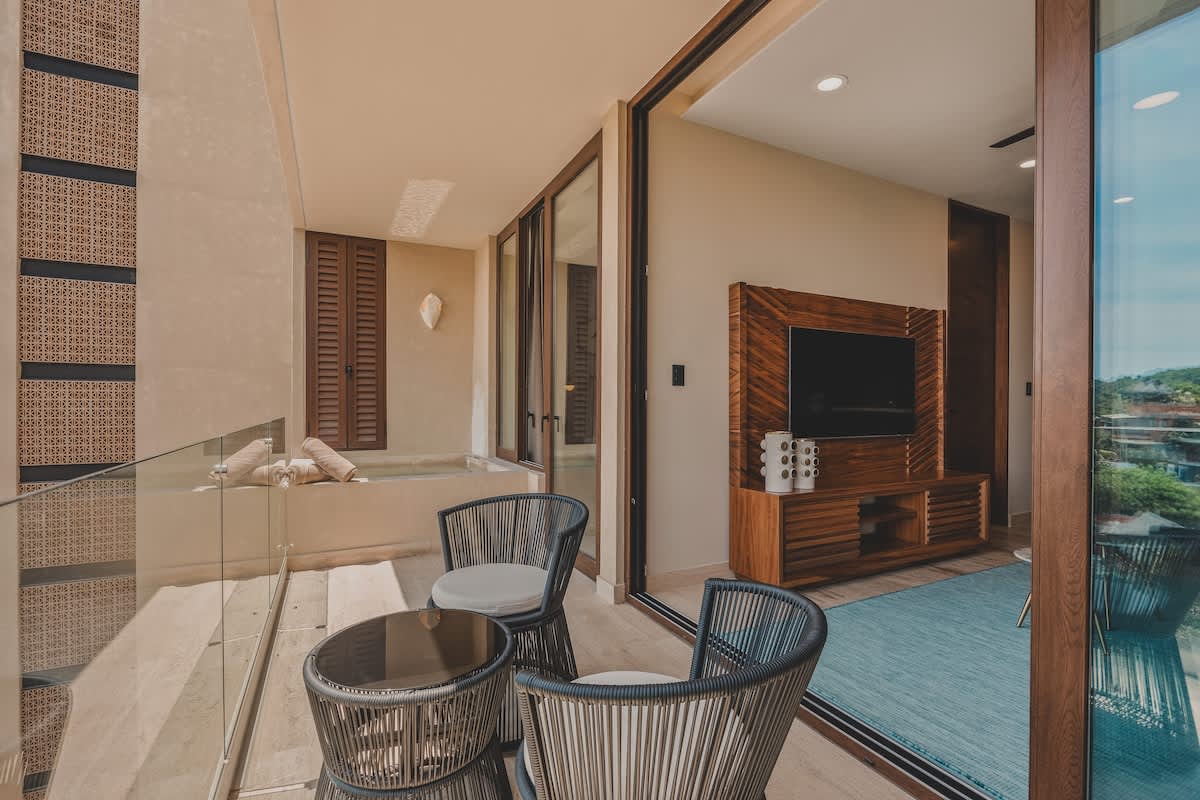 A spacious balcony combines a seating area with two woven chairs and a table, a built-in plunge pool, and large sliding glass doors leading into a bedroom with a wooden media console.