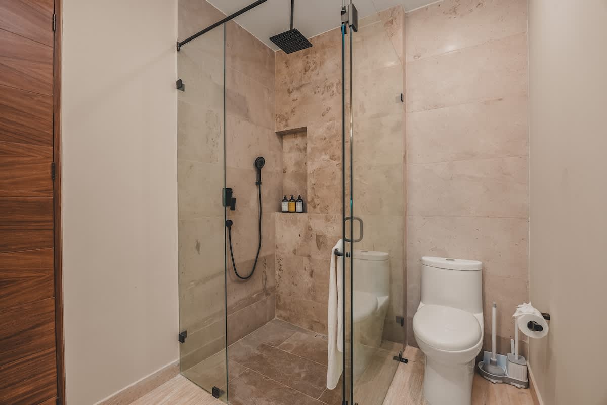 A modern bathroom featuring a glass-enclosed walk-in shower with stone tiles and a black rain showerhead, alongside a white toilet with a toilet paper holder.