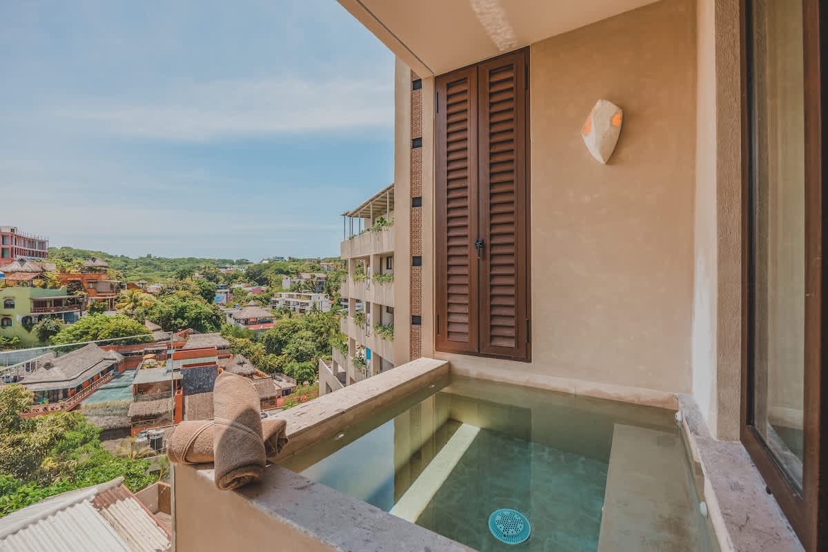 An outdoor plunge pool or hot tub on a balcony, with rolled towels on the edge, overlooking a lush green landscape and distant buildings under a clear sky.