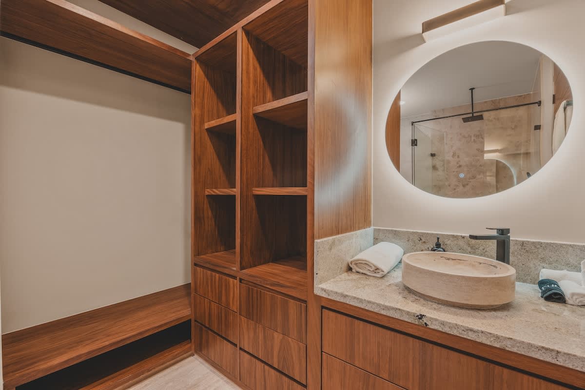 A stylish bathroom vanity area with a light-colored stone countertop, a unique oval stone vessel sink, and a large illuminated round mirror, integrated with an open wooden closet system.