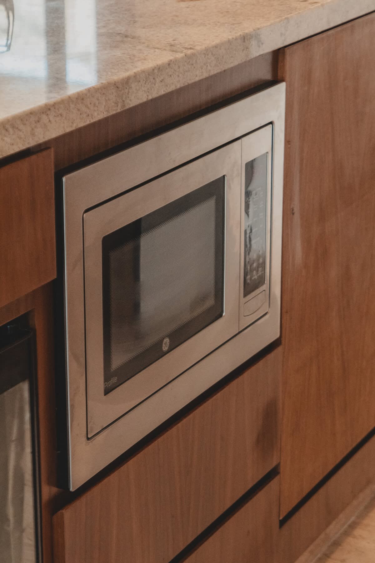 A built-in stainless steel GE Profile microwave is seamlessly integrated into warm-toned wooden cabinetry, set beneath a speckled granite countertop. The sleek design provides a modern and functional kitchen appliance.
