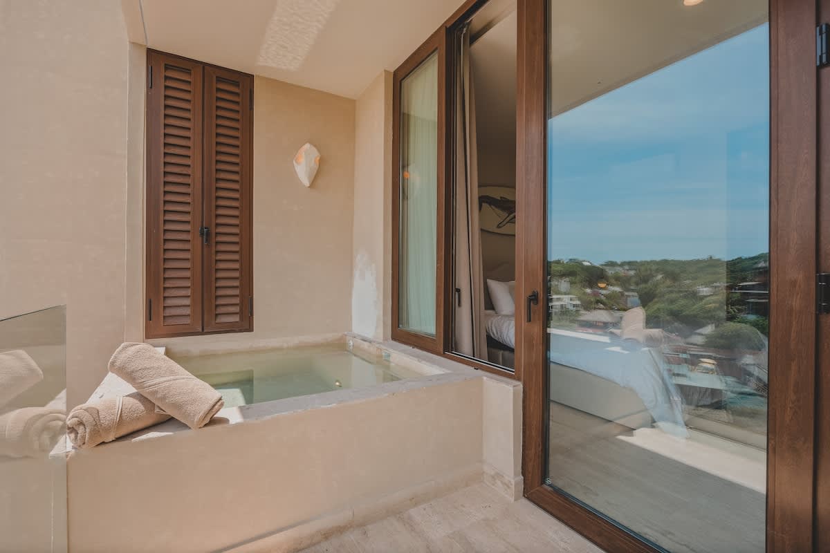 A private balcony features a built-in plunge pool or hot tub with stacked towels, adjacent to a bedroom visible through large sliding glass doors, offering a bright outdoor space.