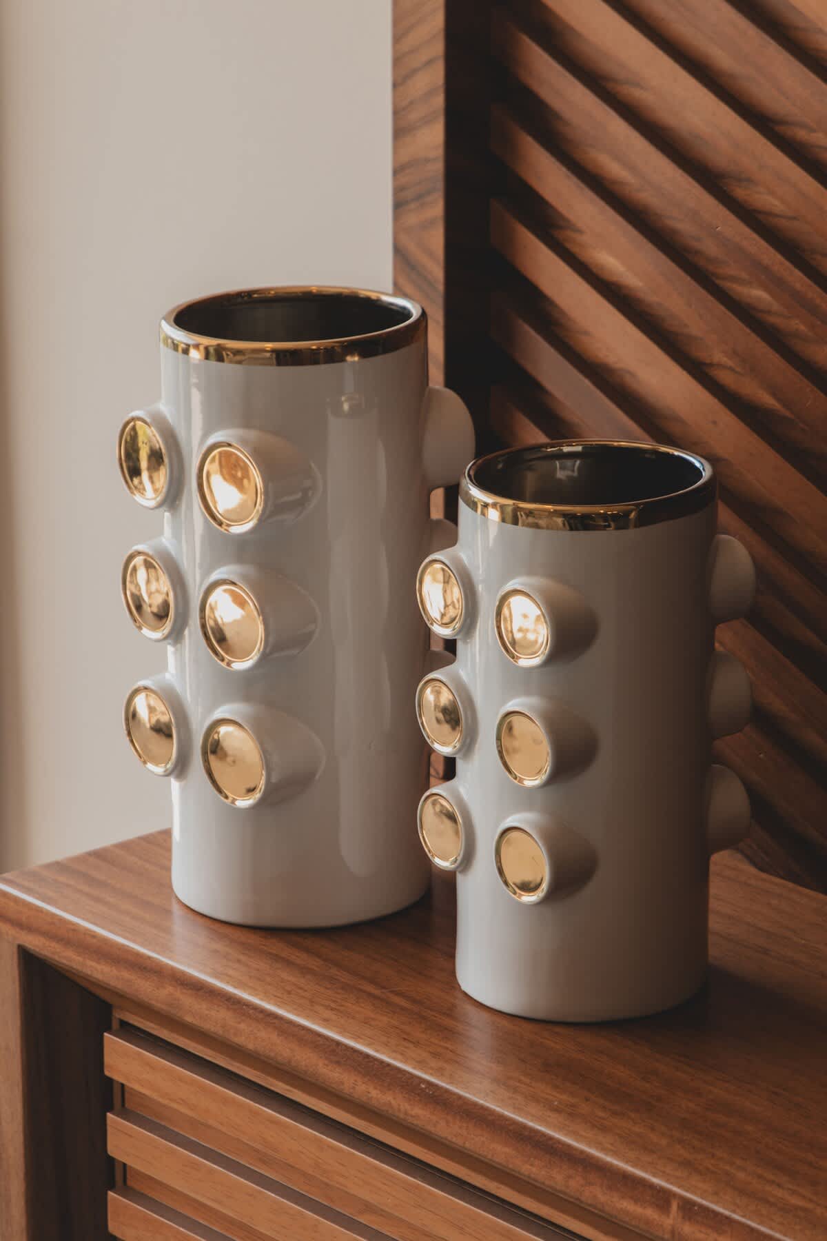 Two decorative white ceramic vases with gold accents and a textured, studded design are displayed on a polished wooden surface. The unique and modern aesthetic adds a touch of elegance to the interior decor.