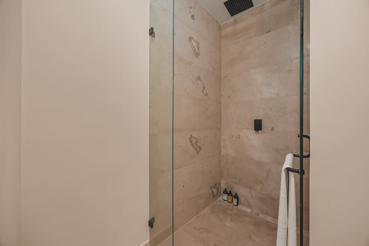 A modern walk-in shower with light-colored stone tile walls and floor, featuring a black rain showerhead and a handheld shower, enclosed by clear glass panels.