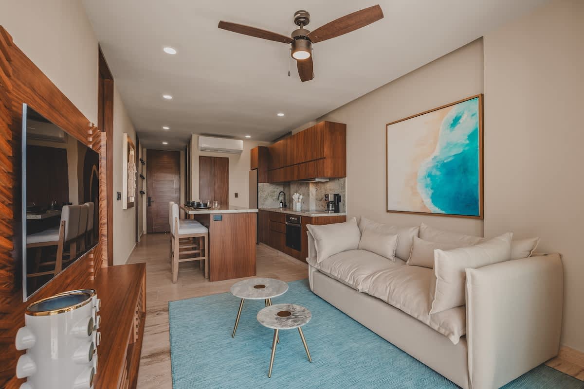 A modern open-concept living room features a light-colored sofa, a blue area rug, and two marble-top coffee tables, flowing into a kitchen with dark wood cabinetry and a breakfast bar.