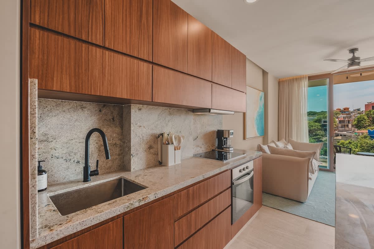Modern kitchen with rich wood cabinetry, light granite countertops, and a matching backsplash. It features a black gooseneck faucet over a stainless steel sink, an induction cooktop, and a coffee maker, with a living area visible through a glass door in the background.