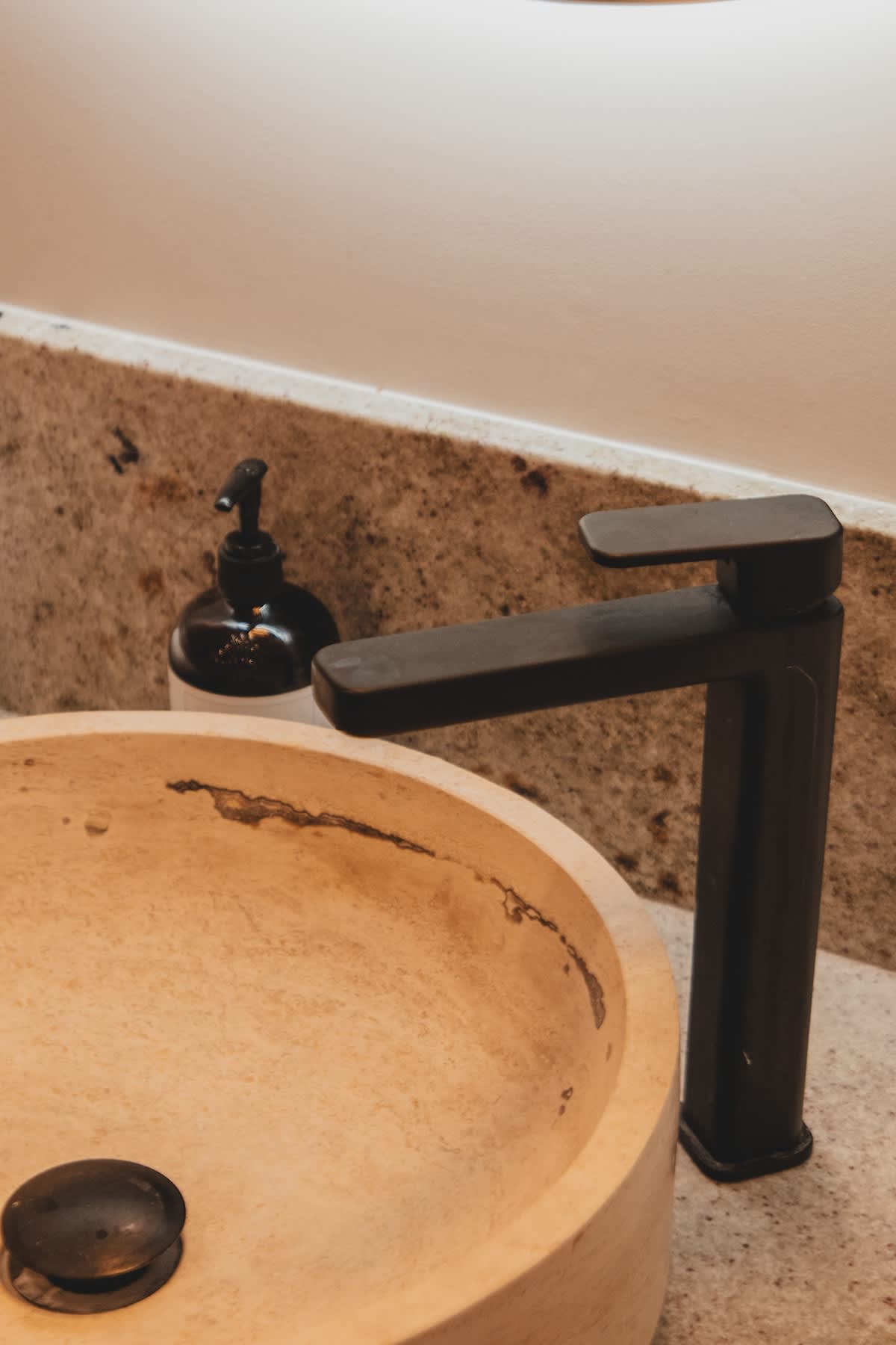 A modern black faucet stands over a rustic, light-colored stone vessel sink, set against a speckled granite countertop and backsplash, with a black soap dispenser nearby.