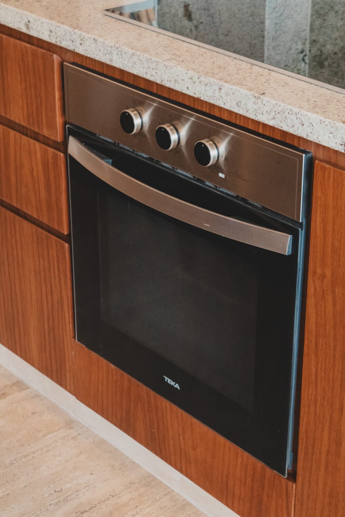 A modern stainless steel oven with a large glass door and three control knobs is built into rich wooden cabinetry, topped by a speckled granite countertop. The sleek design offers a contemporary cooking solution.