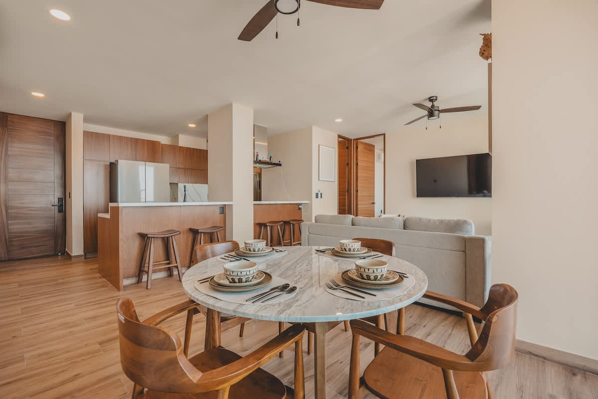 Elegant dining area with a round marble-top table and four wooden chairs, seamlessly flowing into an open-concept kitchen with a breakfast bar and a living room beyond.