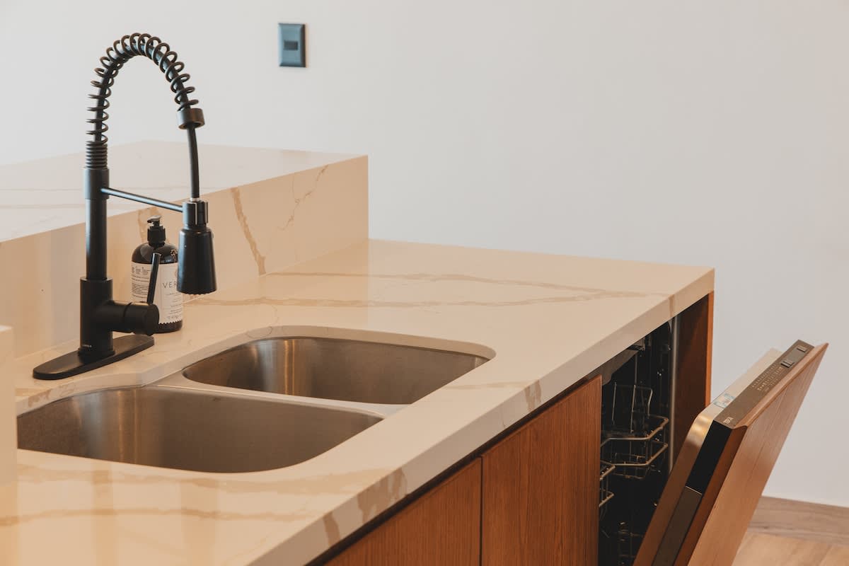 A contemporary kitchen area highlights a double stainless steel sink with a black pull-down faucet, integrated into a light quartz countertop with brown veining. An open dishwasher, seamlessly built into the wooden cabinetry, reveals its racks.