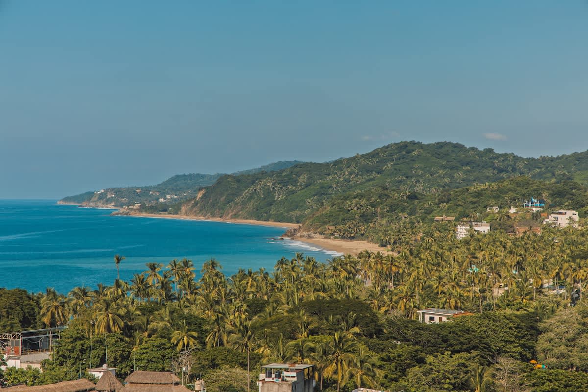 Panoramic aerial view of a lush green coastline with dense palm trees, white sandy beaches, and the clear blue ocean stretching to the horizon under a bright sky.